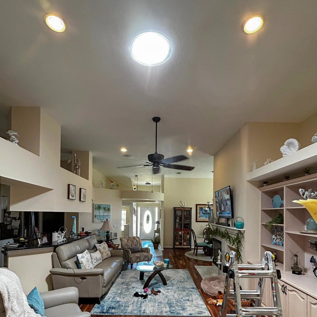 Spacious living room with beige walls and high ceiling, featuring a Sun Tunnel skylight, ceiling fan, leather sofa, TV, decorative shelves, and a ladder. Cozy ambiance.