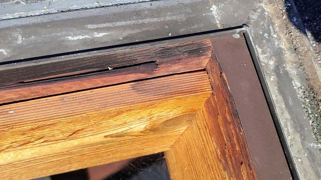 Close-up of a worn wooden skylight frame corner with visible peeling and weathered wood, indicating water damage.