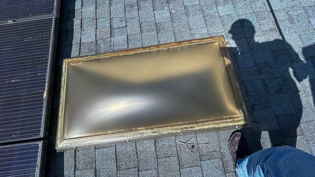 A damaged, discolored skylight on a shingled roof next to solar panels. A person's shadow and feet, wearing jeans, are visible near the skylight.