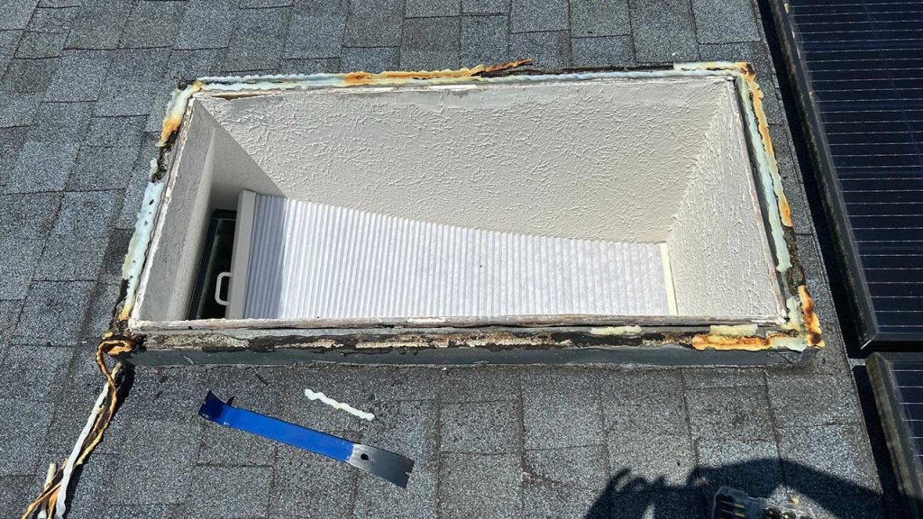 Rectangular skylight frame on a shingled roof, missing the glass. Exposed insulation surrounds the opening. A tool rests nearby, suggesting repairs.