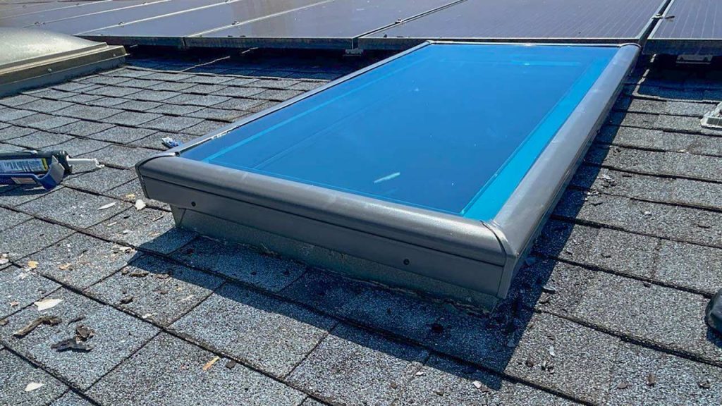 A blue solar skylight is installed on a shingled roof, surrounded by roofing materials and tools. Nearby, solar panels are visible, indicating a renewable energy setup.