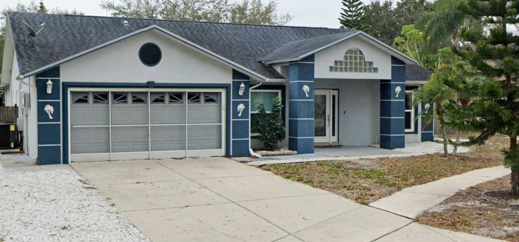Suburban house with a dark roof and blue accents, featuring a two-car garage and palm tree decor. Surrounded by a driveway and sparse greenery.